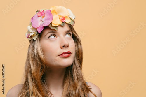 Portrait of a young blonde girl with a headband of flowers and plants on her head looking upwards on a yellow background. summer and nature concept. copy space