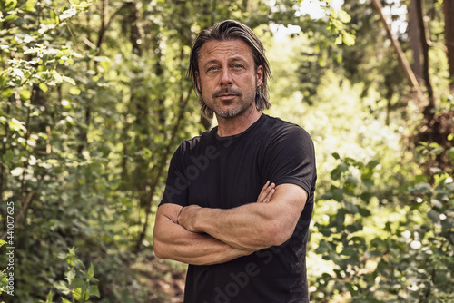 Blond man in a black t-shirt with crossed arms standing in a sunny summer forest.