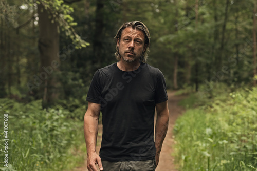 Blond man in a black t-shirt walks on a path in a lush forest.