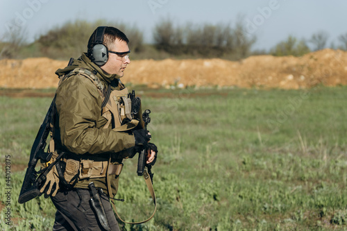 armed young white man in military uniform wearing headphones and glasses at the training ground