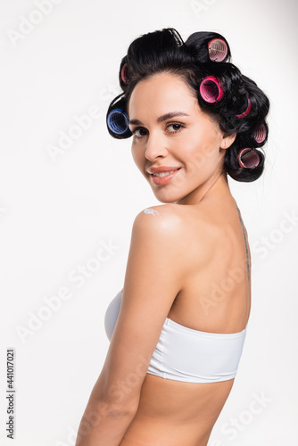 smiling young woman in curlers looking at camera with applied cream on shoulder isolated on white.