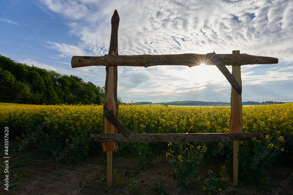 Fototapeta premium Standing wooden window frame in front of yellow rape field. Green forest on the side. Sun star against gray cloudy sky.