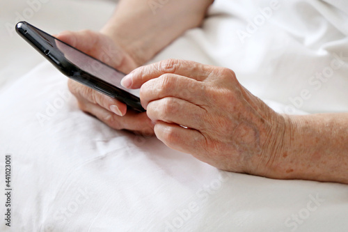 Elderly woman with smartphone in a bed, mobile phone in wrinkled female hands close up. Concept of online communication in retirement, sms, social media