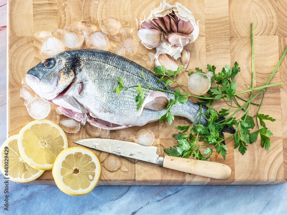 Fresh fish Dorado. Raw sea bream with parsley, onion, garlic and lemon slices covered with ice ready to cook on wooden board. Top view. Marble background. Fresh fish Dorado. Raw sea bream with parsley, onion, garlic and lemon slices covered with ice ready to cook on wooden board. Top view. Marble background.