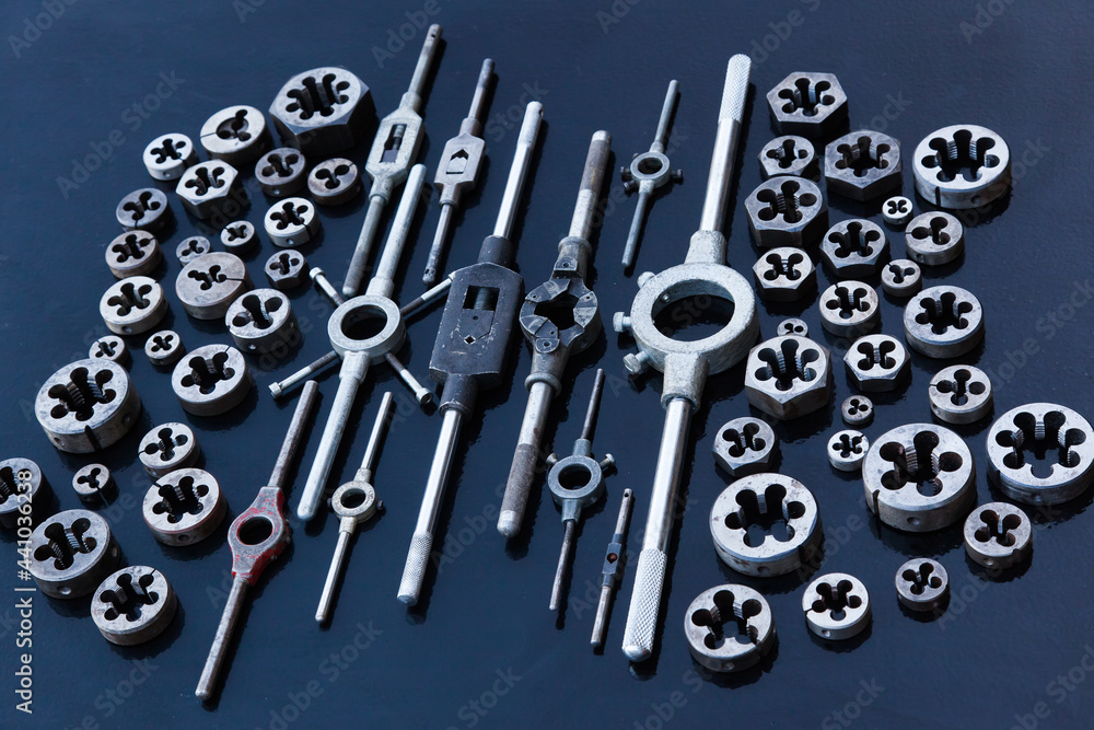 A lot of dies and holders for them of different sizes for threading in metal, on a black background.