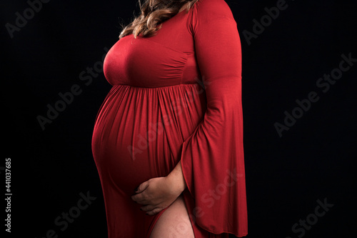 Unrecognizable pregnant woman wearing a long red dress is hugging her big belly while expecting