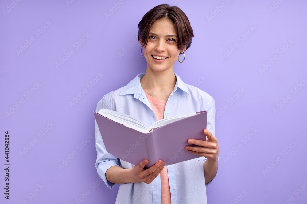 Confident female reading book, isolated on purple background. Young woman in casual wear looking at camera and smiling, holding book in hands Confident female reading book, isolated on purple background. Young woman in casual wear looking at camera and smiling, holding book in hands