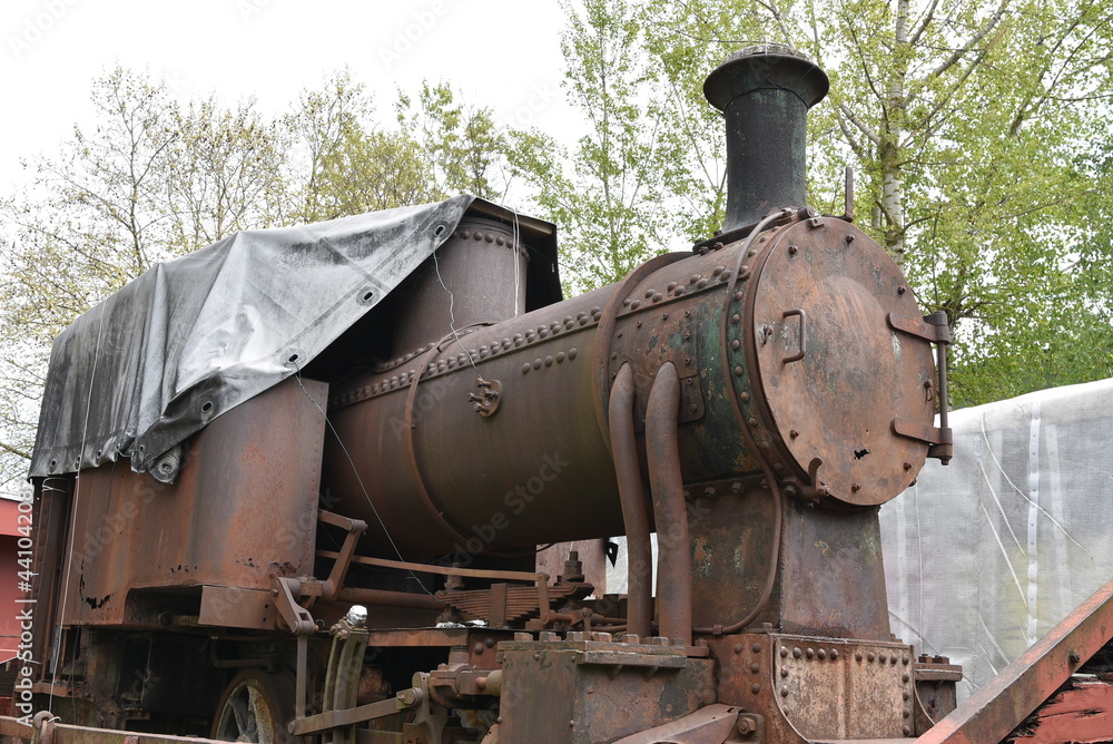Front-end view on discarded rusty freight steam locomotive loaded on old wagon which is standing on blind railroad tracks in abandoned industrial zone. Cabin si covered with tarpaulin. Front-end view on discarded rusty freight steam locomotive loaded on old wagon which is standing on blind railroad tracks in abandoned industrial zone. Cabin si covered with tarpaulin.