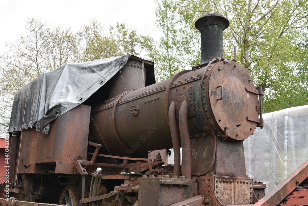 Front-end view on discarded rusty freight steam locomotive loaded on old wagon which is standing on blind railroad tracks in abandoned industrial zone. Cabin si covered with tarpaulin.