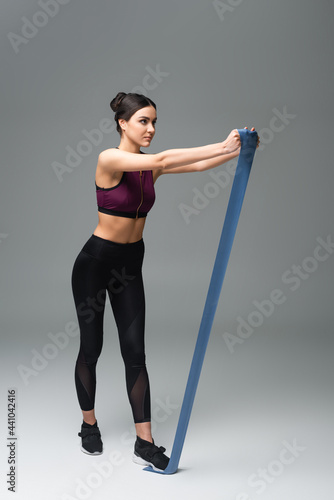 full length view of woman in black sportswear exercising with elastics on grey background.