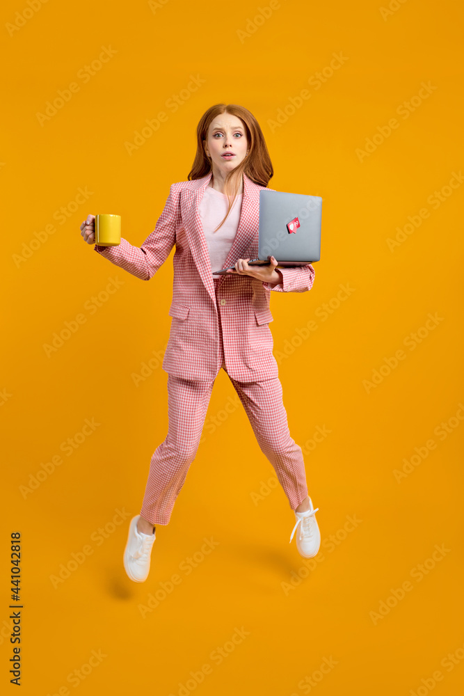 Redhead woman jumping going using laptop web project isolated over yellow background in studio, holding cup of coffee in hands, miss the deadline, hurry up. Full-length portrait Redhead woman jumping going using laptop web project isolated over yellow background in studio, holding cup of coffee in hands, miss the deadline, hurry up. Full-length portrait