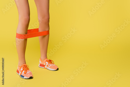 cropped view of sportswoman training with resistance band on legs on yellow background.
