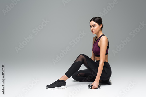 young sportswoman looking away while sitting with dumbbell on grey background.