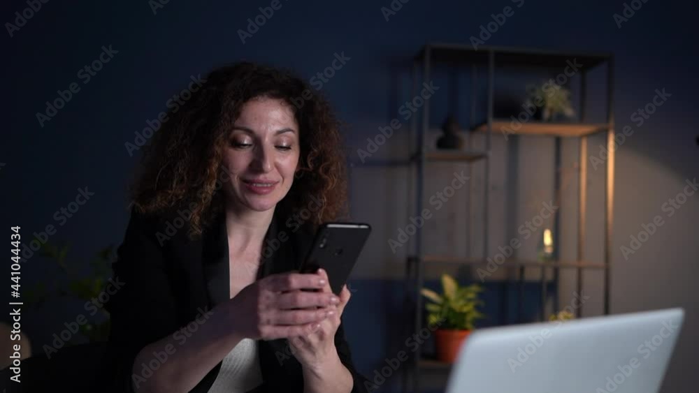 Overworking girl working in the office late at night. Attractive curly-haired business woman is typing text on a laptop keyboard and smiling Overworking girl working in the office late at night. Attractive curly-haired business woman is typing text on a laptop keyboard and smiling