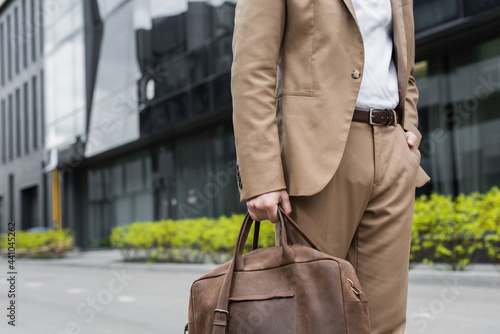 cropped view of businessman in suit holding leather bag and standing with hand in pocket on urban street.