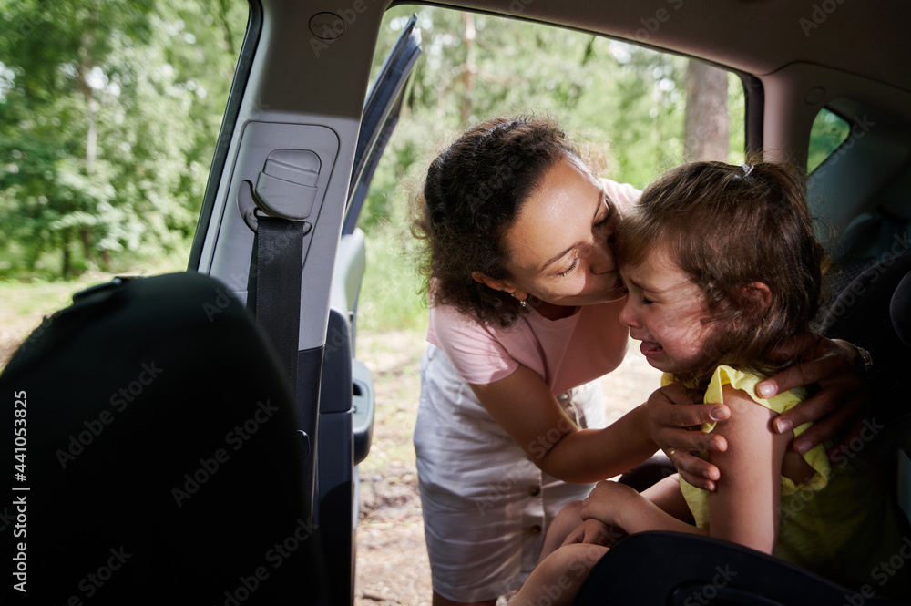 Mom consoles her crying baby girl, who doesn't want to sit in a car seat. Woman seats the child in the child's car seat