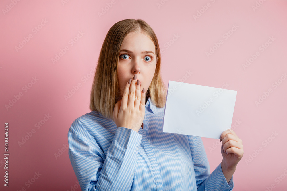 Emotional portrait of surprised young woman show empty mockup copy space white card. Blonde woman In shirt cover mouth with hand in surprise with blank board for text over color pink background. Emotional portrait of surprised young woman show empty mockup copy space white card. Blonde woman In shirt cover mouth with hand in surprise with blank board for text over color pink background.