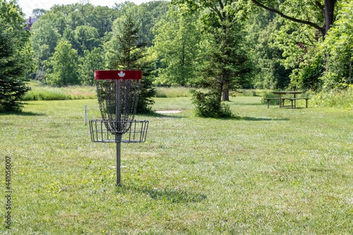 Empty red disc golf basket with white Canadian maple leaf on the top. Metal chains. Basket is on grass of disc golf course, trees in the background.