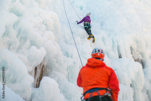 Man standing and controlling a safety top rope while female with ice climbing equipment, climbing on a frozen waterfall photo