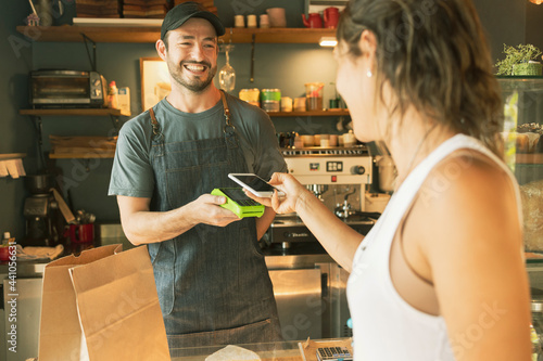 Beautiful young woman paying for purchases with her smartphone in a Brazilian organic artisanal bakery. Small business and technology concept.