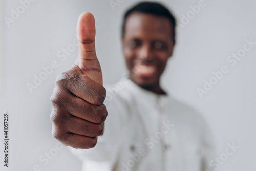 Happy African-American blurry person in white shirt shows thumb up gesture posing for camera on light grey background extreme closeup