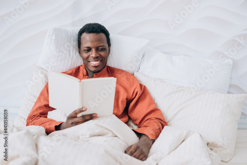 Handsome black young man in pajamas reads interesting book lying in comfortable bed with soft pillows and blanket in room