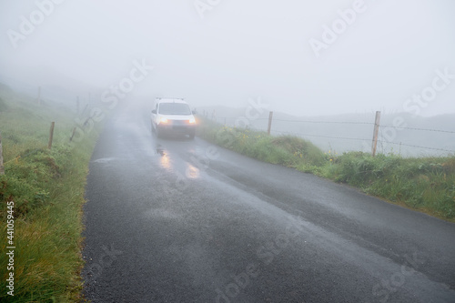 Fog over small country asphalt road. Car with lights out of focus in the mist. Selective focus, dangerous driving conditions concept