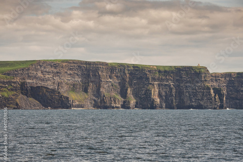 View on Cliff of Moher from a board of a ferry or cruise ship. West coast of Ireland. Popular tourist destination. National landmark. Cloudy sky. Calm surface of Atlantic ocean.