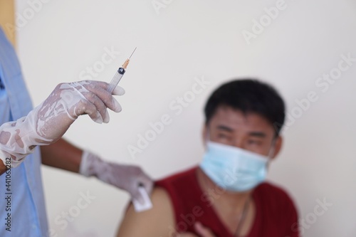 Concept : Covid-19 vaccination. Close-up syringe of vaccine in doctor's hand before injection. Blurred photo of man wears hygienic face mask background. Feeling afraid of injection. Fear, hesitated.
