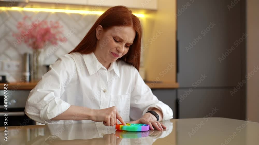 Stressed redhead young woman playing with fidget sensory toy to relieve stress to simple way to be in calm and harmony at home. Negative redhead lady playing with antistress popit toy sitting at table Stressed redhead young woman playing with fidget sensory toy to relieve stress to simple way to be in calm and harmony at home. Negative redhead lady playing with antistress popit toy sitting at table