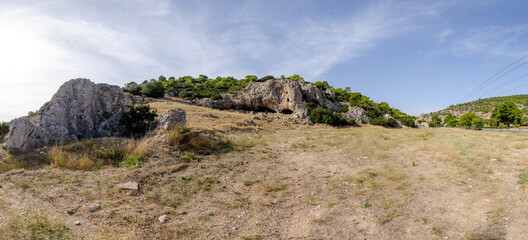 Panoramic shot of a dirt field leading up to rocky peak with forest and blue sky in the background