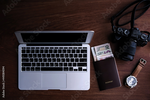 top view of traveling photographer with mobile laptop, passport with many banknotes, digital camera, and wrist watch put together on dark red wooden table with focusing only on notebook computer