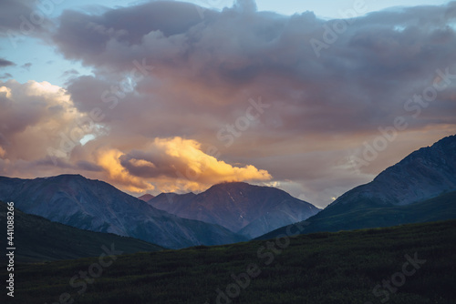 Beautiful mountain scenery with golden dawn light in cloudy sky. Scenic mountain landscape with illuminating color in sunset sky. Silhouettes of mountains on sunrise. Gold illuminating sunlight in sky