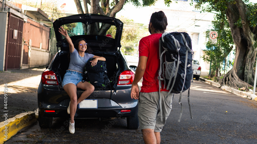 Obraz premium Young woman sitting in the trunk of the car next to her suitcase waving to the friend who is arriving with his suitcase, ready to go on a trip