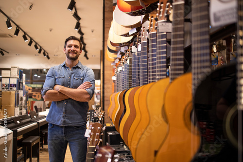 Portrait of young talented musician standing in music shop by large collection of guitar instruments.