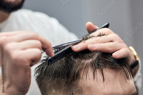 Professional barber making haircut to young man using scissors and comb at barbershop. Close-up, selective focus