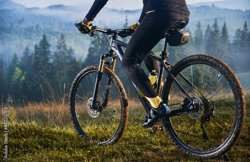 Cropped view of man in cycling suit riding bicycle on grassy hill. View of majestic mountains on the blurred background. Concept of sport, bicycling and nature.
