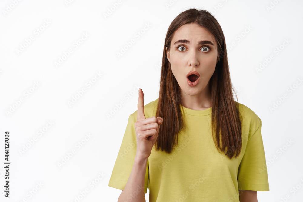 Portrait of shocked girl gasp, pointing finger up and staring in awe, impressed by smth, look with disbelief, standing in t-shirt against white background Portrait of shocked girl gasp, pointing finger up and staring in awe, impressed by smth, look with disbelief, standing in t-shirt against white background