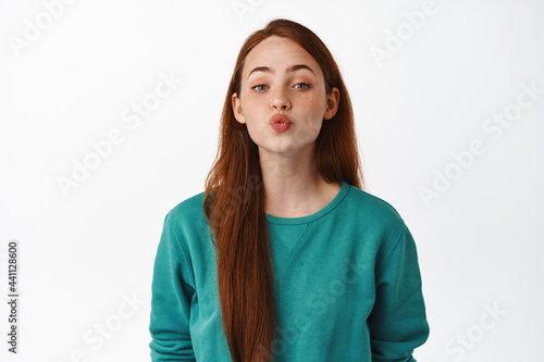Beautiful caucasian redhead girl kissing, pucker lips and leans for kiss, romantic relationship or date, standing flirty against white background © Cookie Studio