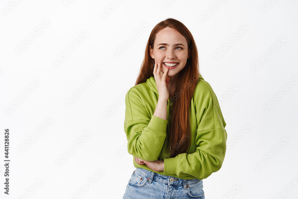 Beautiful ginger girl with pale skin and freckles, looking aside at logo and laughing happy, standing in hoodie and jeans against white background Beautiful ginger girl with pale skin and freckles, looking aside at logo and laughing happy, standing in hoodie and jeans against white background