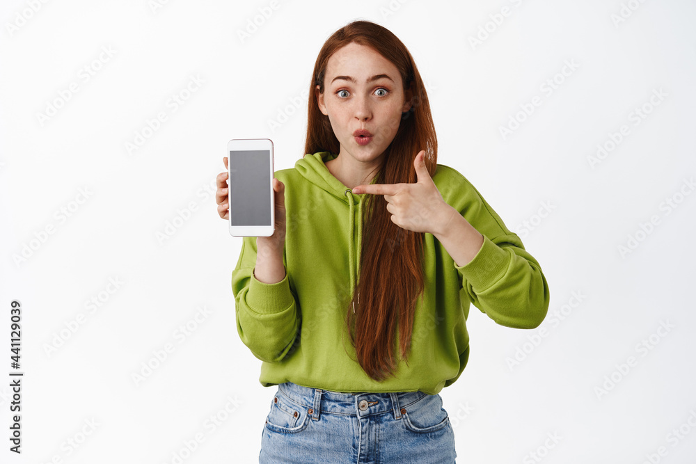 Surprised redhead woman pointing finger at smartphone app, gasping in awe, showing interesting application on mobile phone, white background Surprised redhead woman pointing finger at smartphone app, gasping in awe, showing interesting application on mobile phone, white background