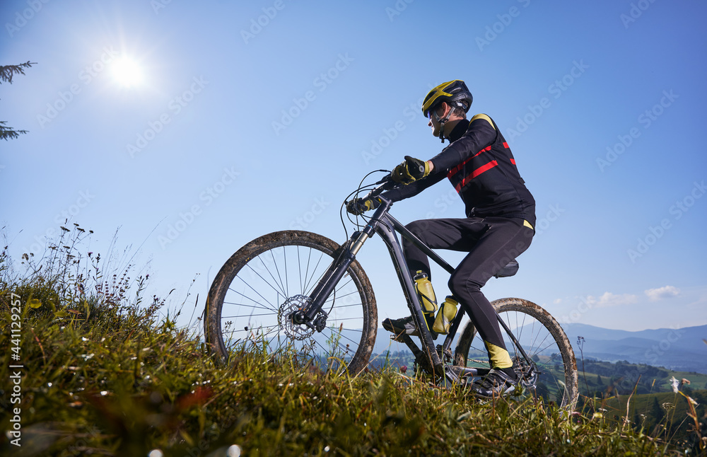 Young man in safety helmet and glasses cycling uphill with blue sky on background. Male bicyclist in cycling suit climbing uphill on mountain bike. Concept of sport, mountain biking and active leisure