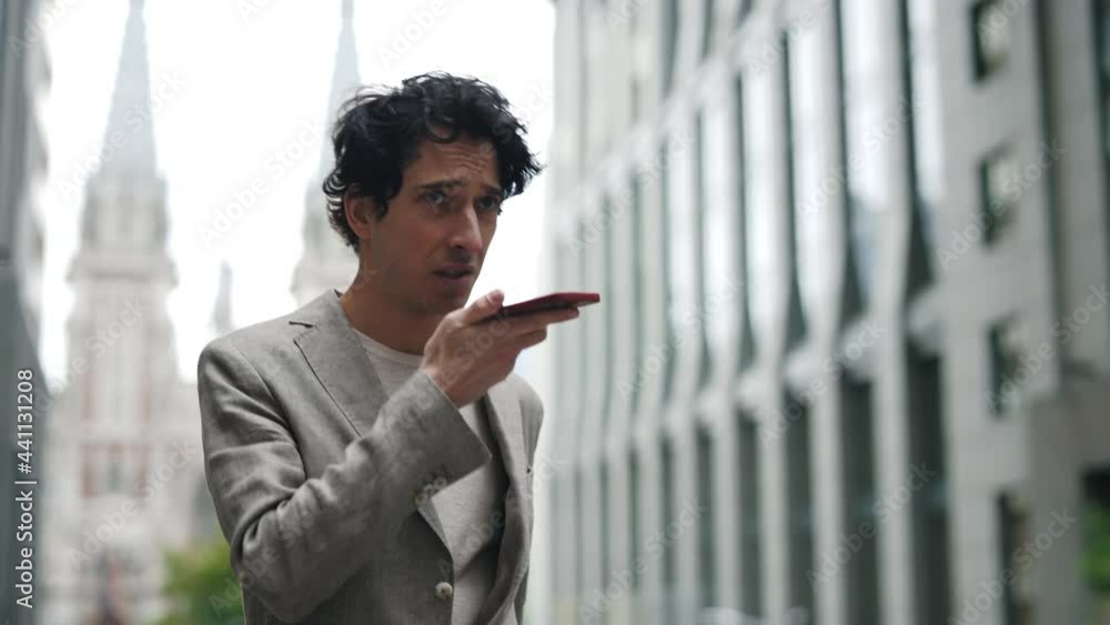Young attractive businessman walking near big modern office building and recording message on his smartphone. Man wearing classical suit is looking satisfied. Business lifestyle/ Young attractive businessman walking near big modern office building and recording message on his smartphone. Man wearing classical suit is looking satisfied. Business lifestyle/