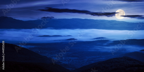 mountainous countryside landscape at night. trees and agricultural fields on hills rolling in to the distant misty valley. ridge beneath a sky in full moon light © Pellinni