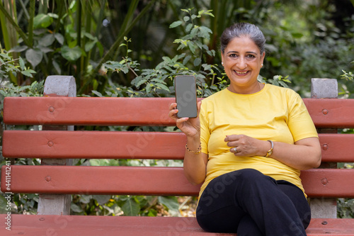 A SENIOR ADULT WOMAN HAPPILY SHOWS HER MOBILE PHONE WHILE SITTING IN A PARK