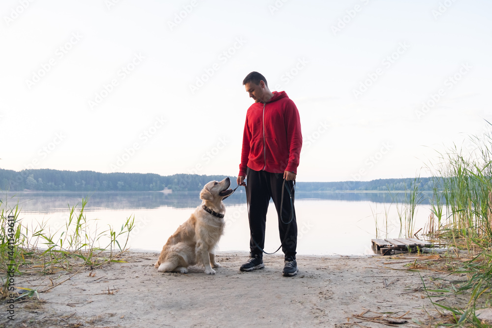 A young man is stroking his dog on the river bank in the dawn sun. A guy and a golden retriever look into the distance on a summer morning. Travel concept with pets.