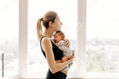 Young mother holding and hugging newborn baby in her arms at home, by the window.