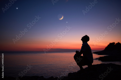 Silhouette of a man looking at the Moon and stars over sea ocean horizon.