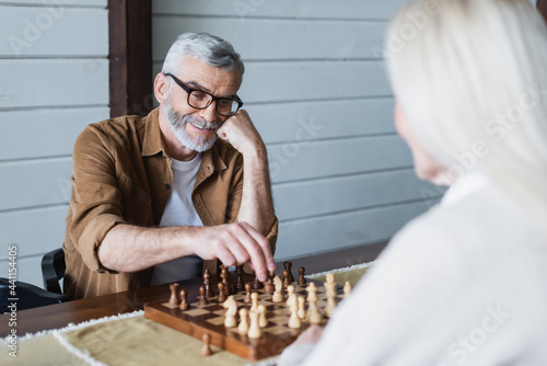 Smiling elderly man playing chess with wife on blurred foreground