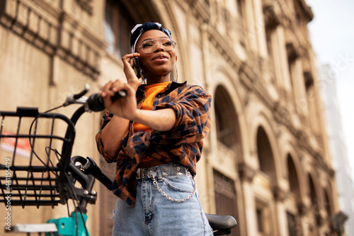 Happy young woman riding bicycle in the city. Beautiful african woman using the phone outdoors.